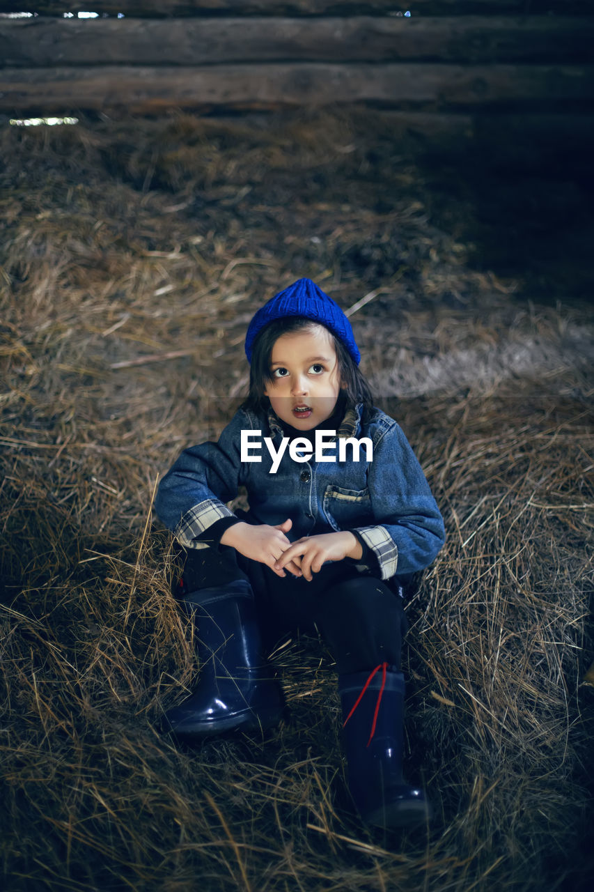 Village boy a child in boots is sitting
on the hay in a barn in warm clothes and a blue hat