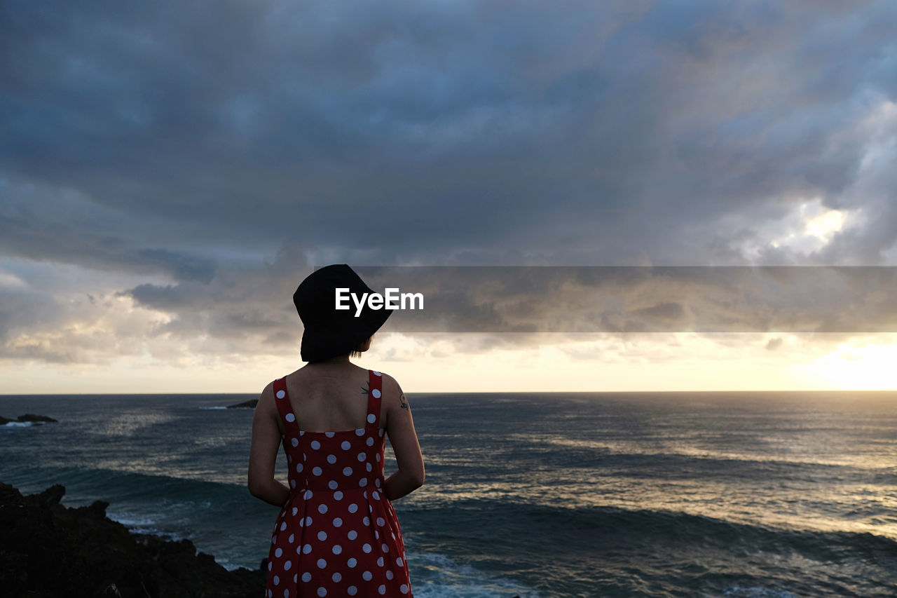 rear view of woman standing at beach against cloudy sky