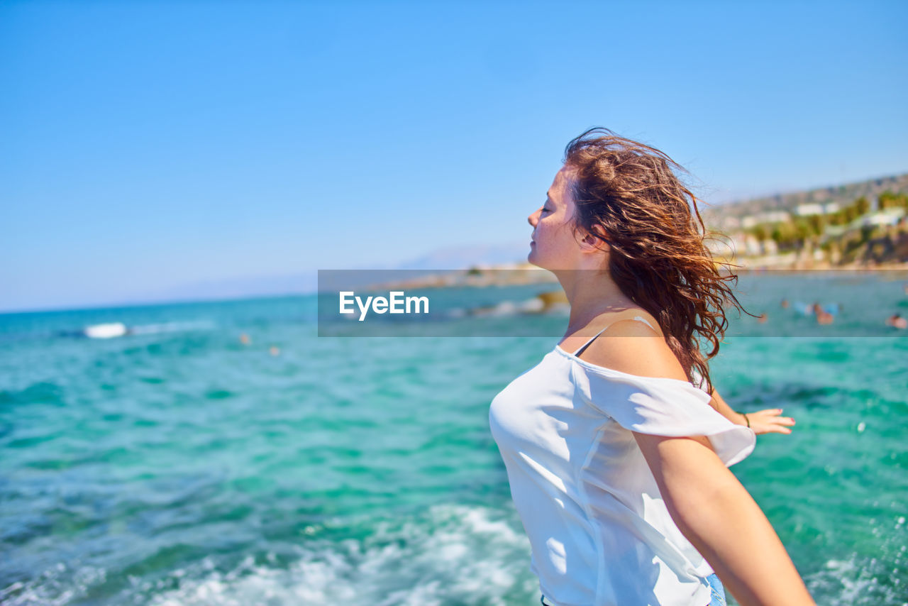 Side view of woman with arms outstretched standing against clear sky at beach