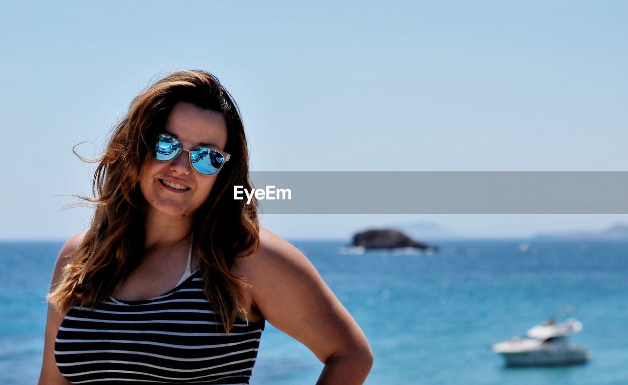 Portrait of smiling young woman standing at beach against clear sky