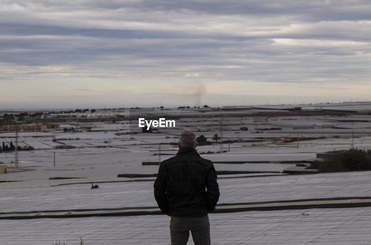 Adult man standing in front of plastic greenhouses against cloudy sky in almeria, spain