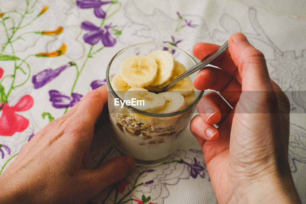 Close-up of a woman's hands holding a glass of yogurt with jam, oat flakes and banana