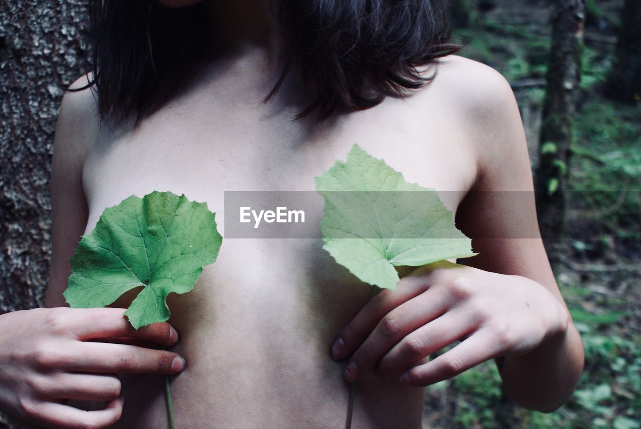 Midsection of shirtless woman covering breast with leaves