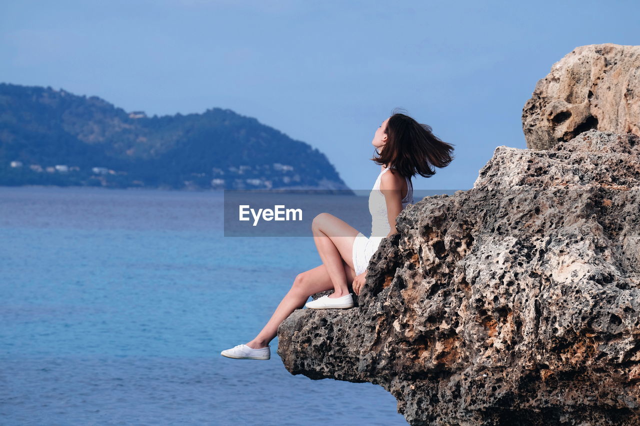 Side view of woman sitting on rock by sea against sky