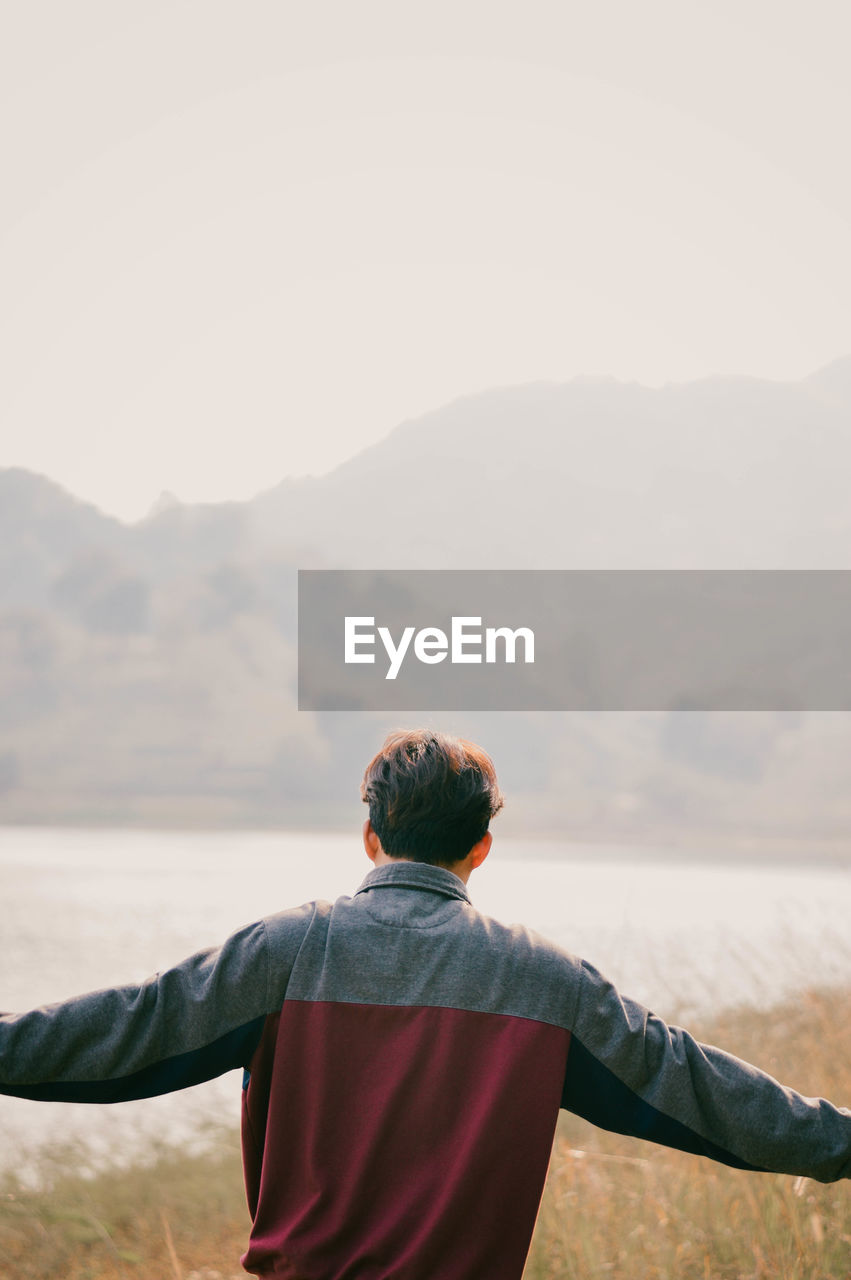 REAR VIEW OF MAN STANDING BY MOUNTAIN