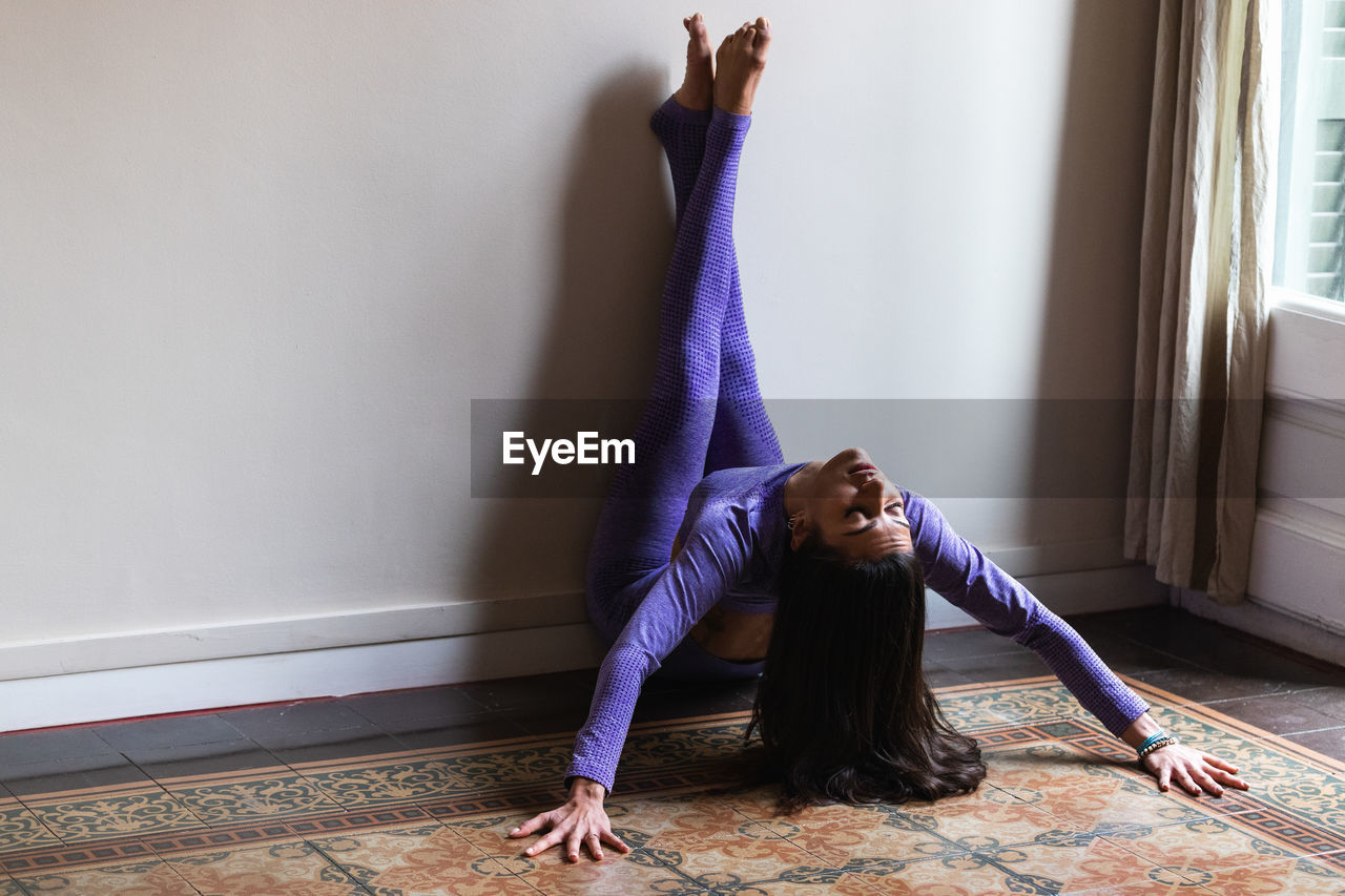 Full body of graceful young hispanic female with long dark hair in sportswear performing asana with raised legs on wall while practicing yoga in cozy apartment