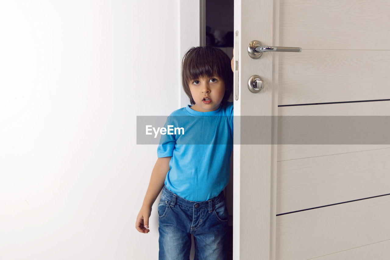 Boy in a blue t-shirt and jeans is standing at home in the white door