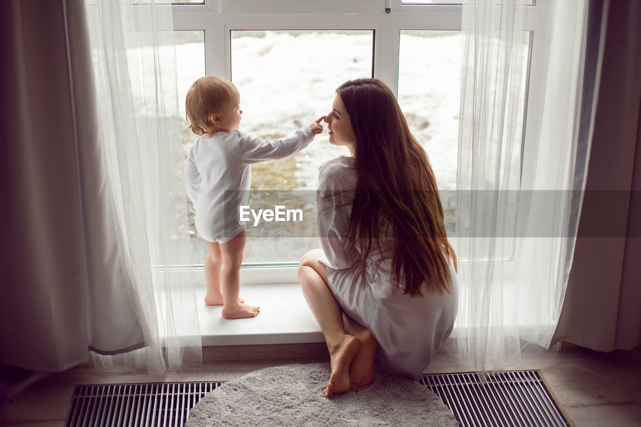 Mother in a white robe sits with a child a blonde daughter at a large window of the house person