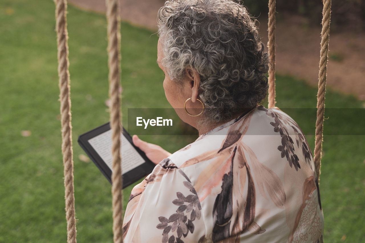 Back view of senior lady wearing blouse sitting in park and reading electronic book
