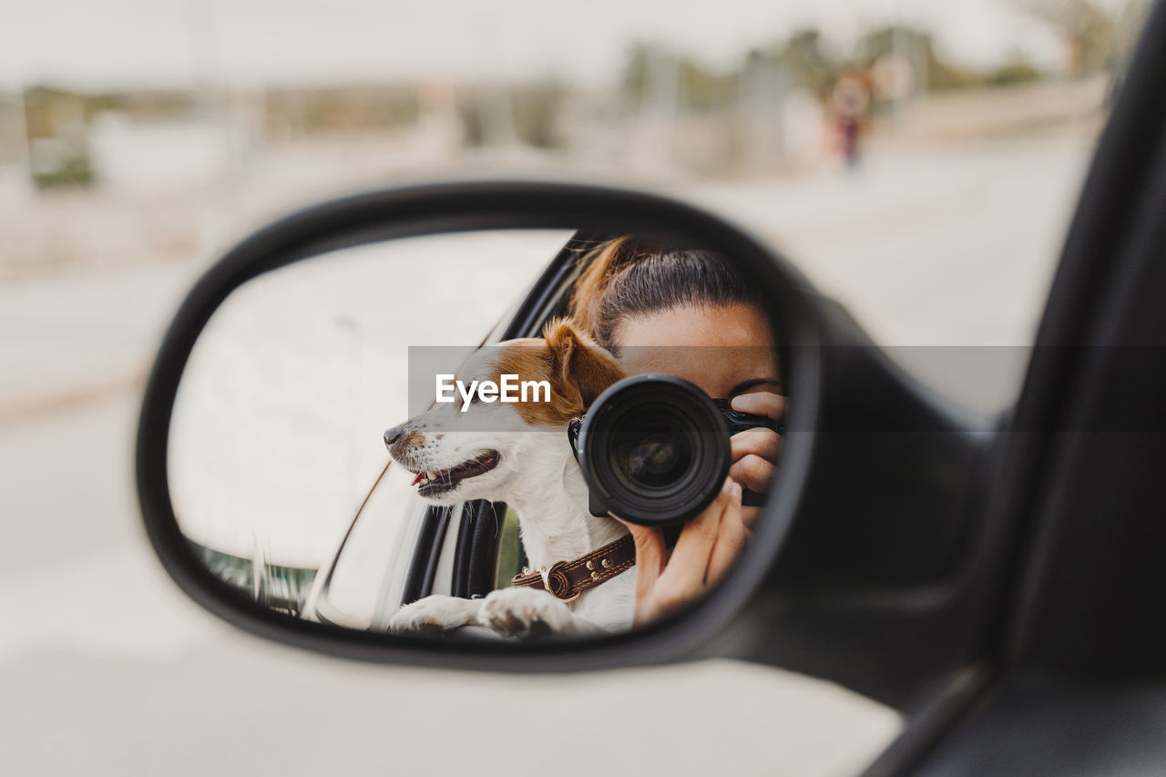 Reflection of woman photographing by dog in side-view mirror car