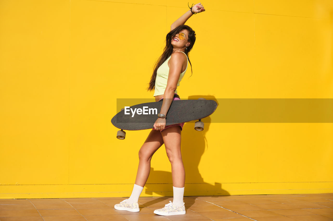 Portrait of young woman standing in front of yellow wall with longboard in hand