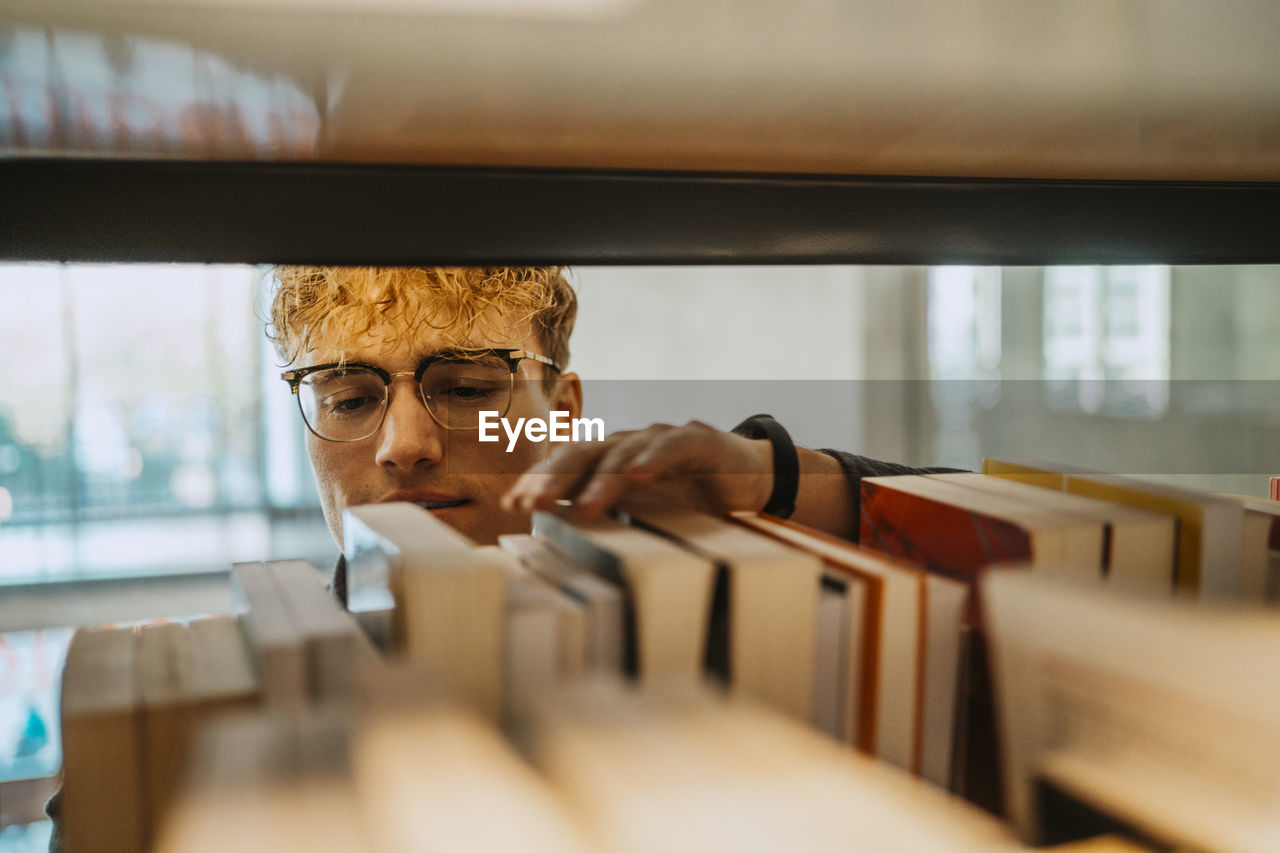 Young man wearing eyeglasses searching book in library at university