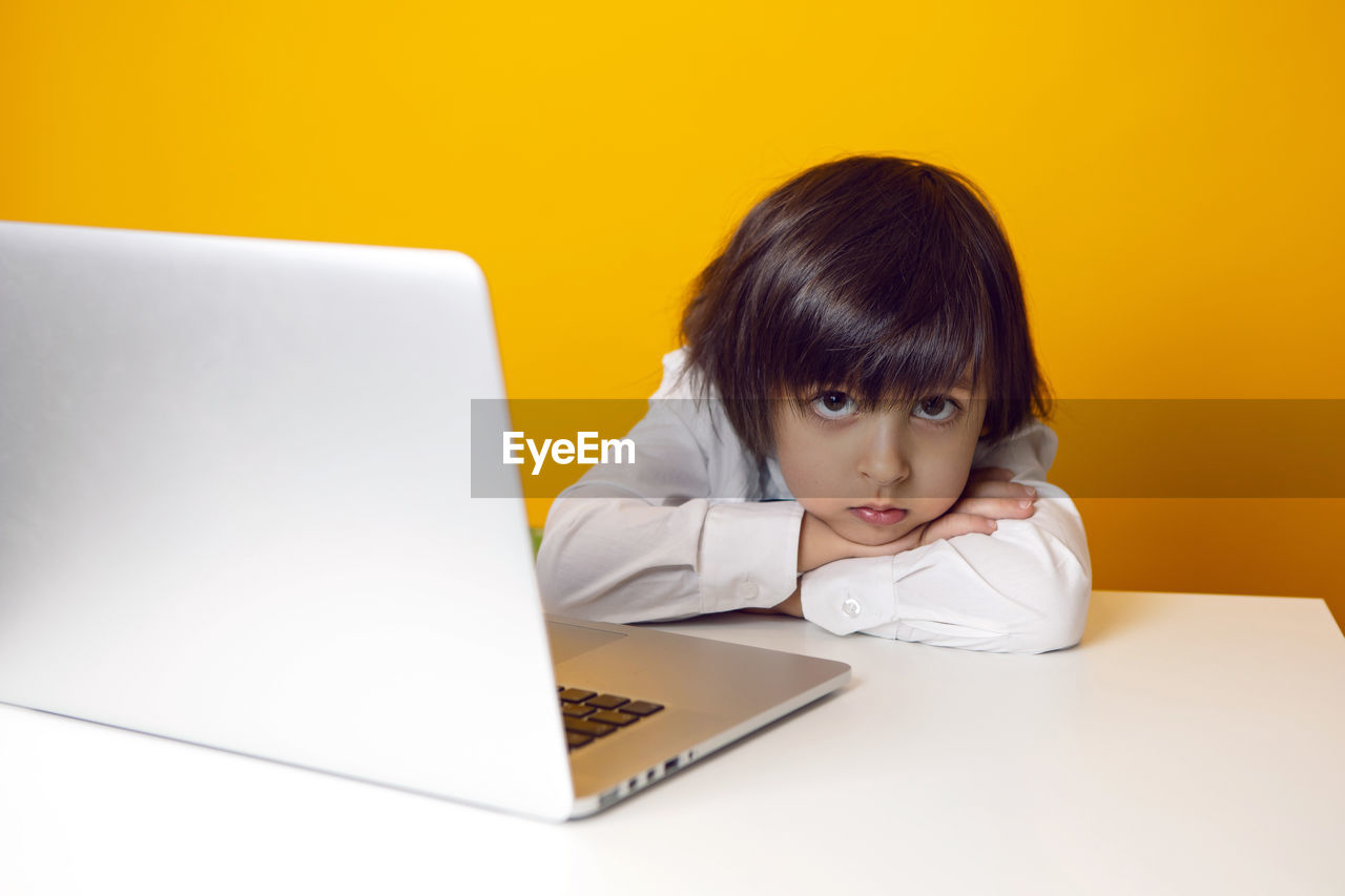 Serious boy child businessman in a white shirt and bow tie sits at a laptop  desk in a yellow office