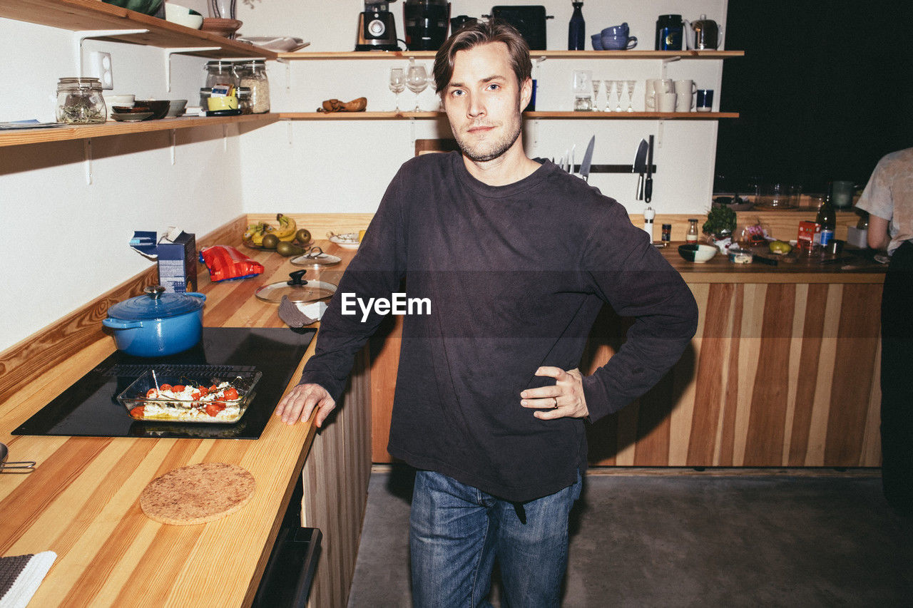Portrait of man standing with hand on hip while preparing food in kitchen at home
