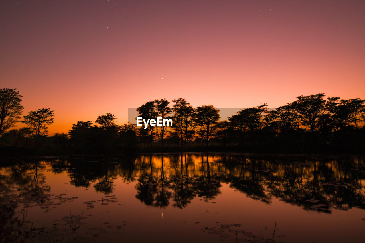 Silhouette trees by lake against orange sky