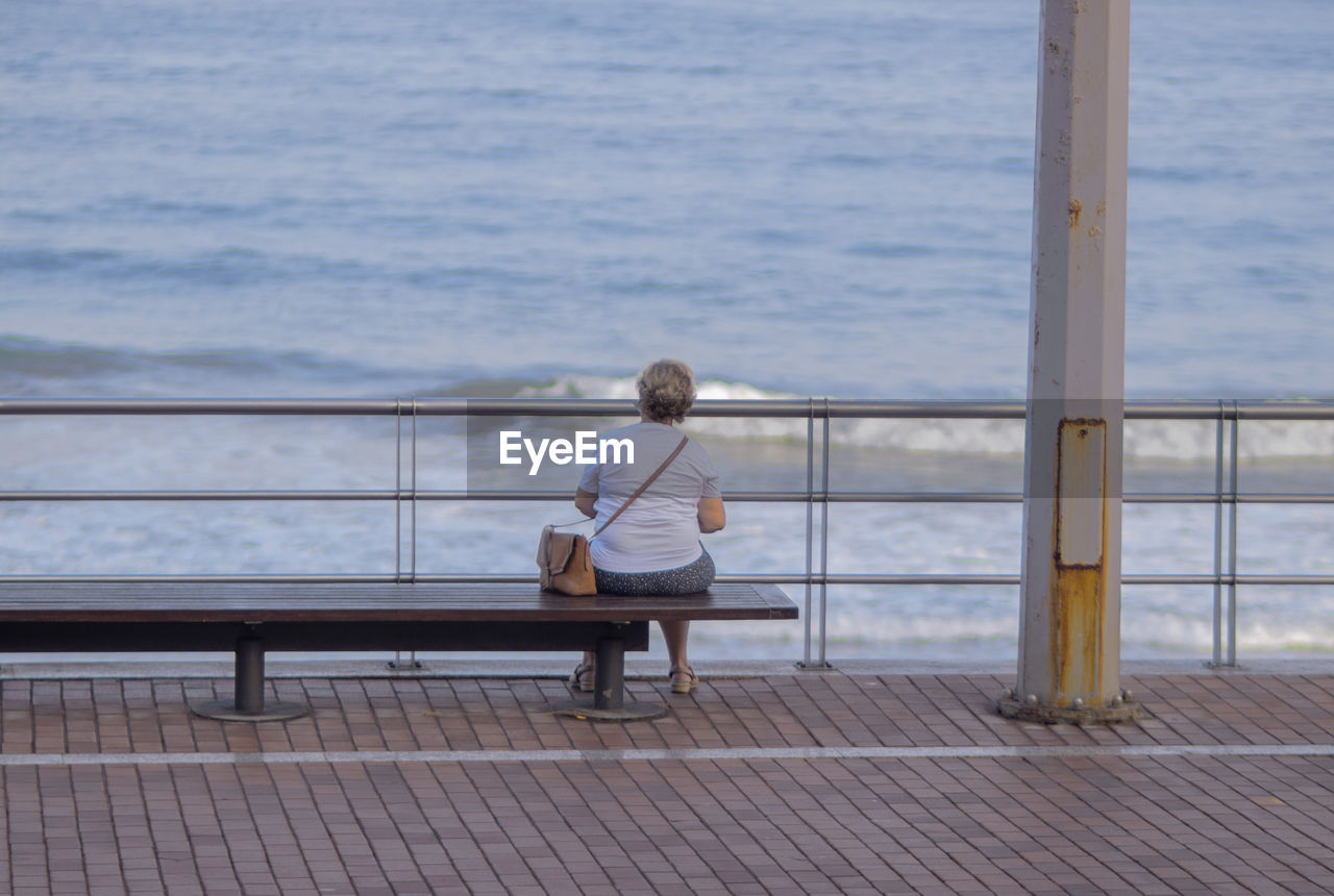 REAR VIEW OF MAN SITTING ON BENCH AT SEA