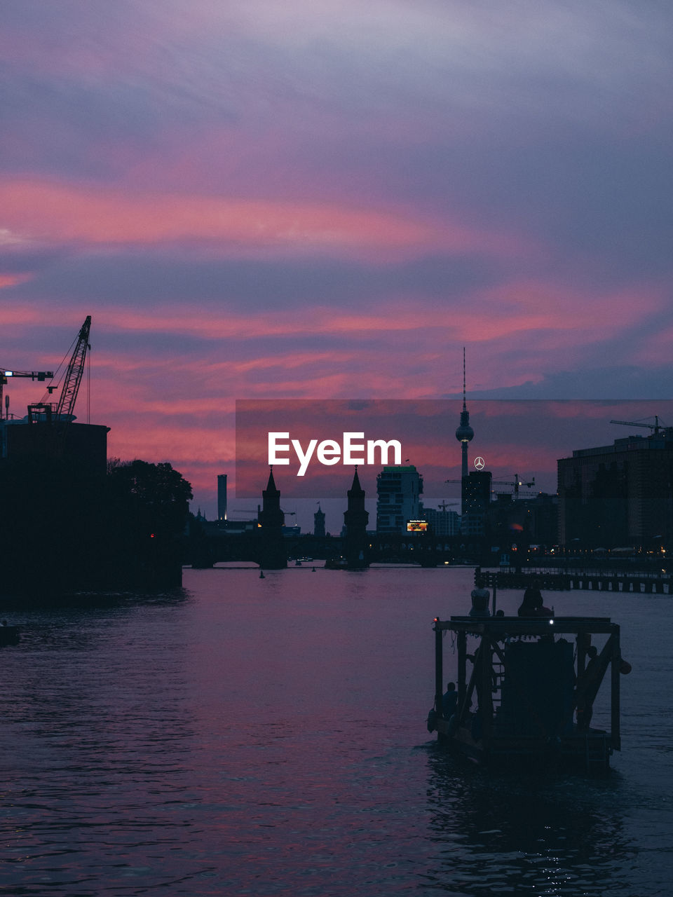 Silhouette buildings by river against cloudy sky at sunset