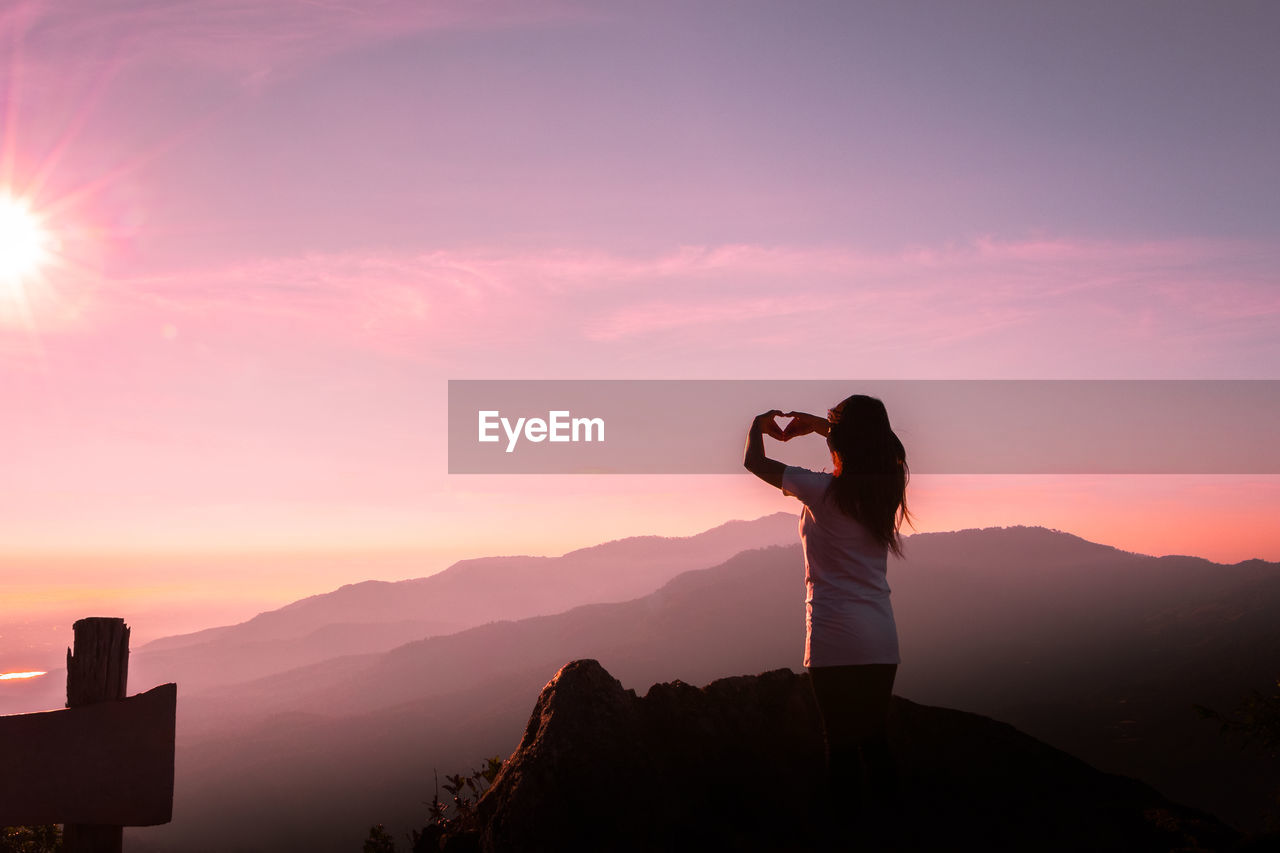 Rear view of woman on mountain making heart shape against sky during sunset