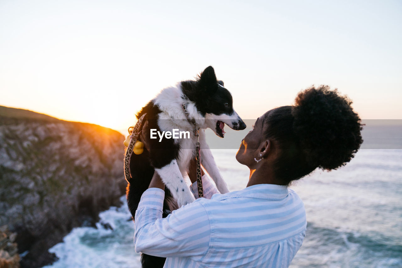 Side view of african american female owner holding cute happy border collie dog while enjoying time together near waving sea at sunset