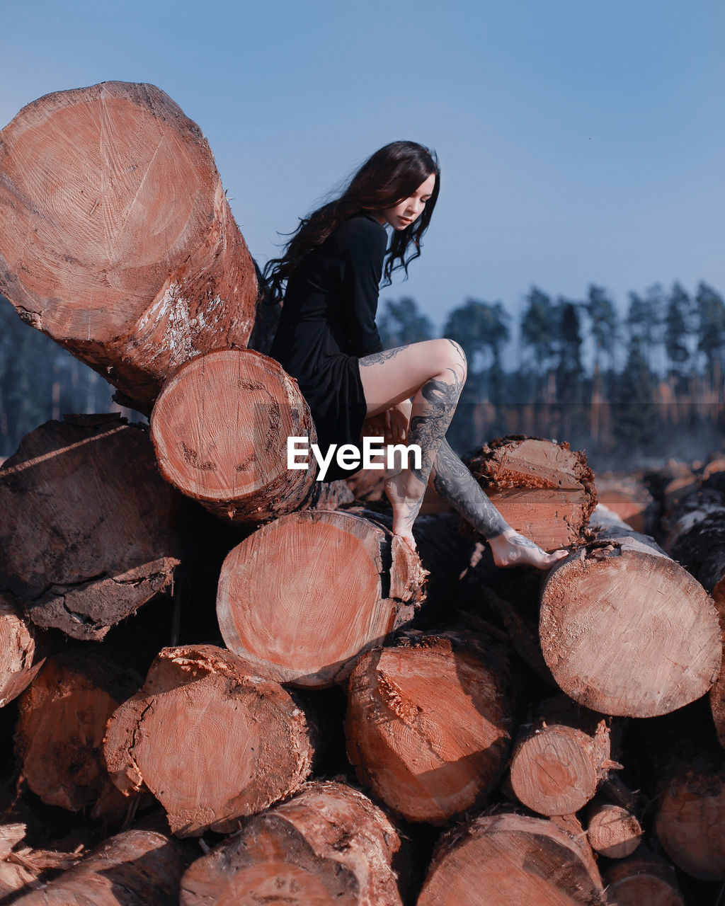 Side view of woman sitting on logs against clear sky