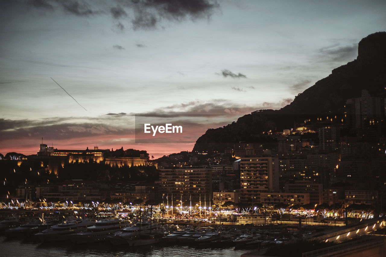 Illuminated buildings by sea against sky at sunset