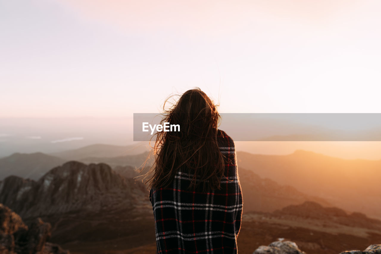 Back view of unrecognizable hiker in blanket standing on stone and observing amazing scenery of highlands valley on sunny day