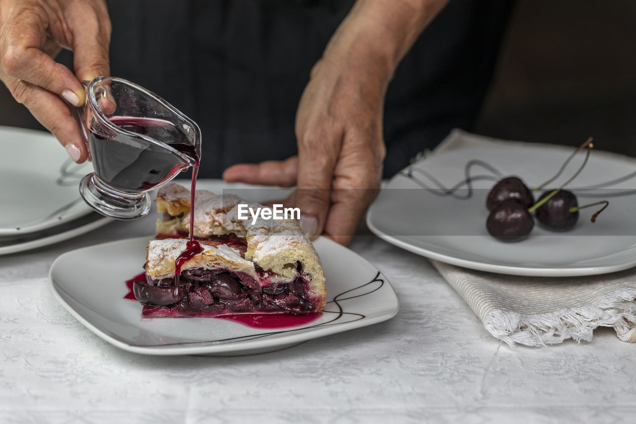 An unrecognisable person pouring red cherry syrup on a slice of cake filled with seasonal cherries.
