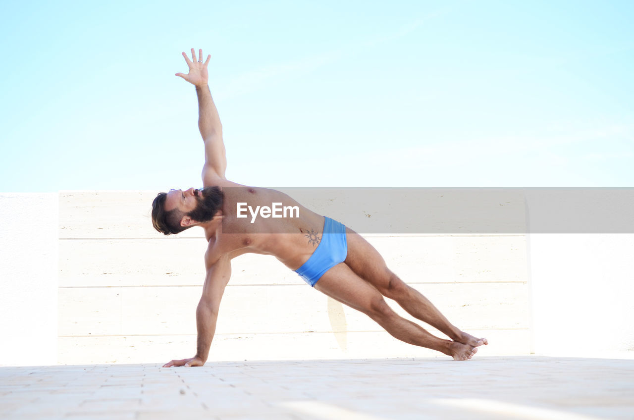 Full length of shirtless man practicing yoga against sky