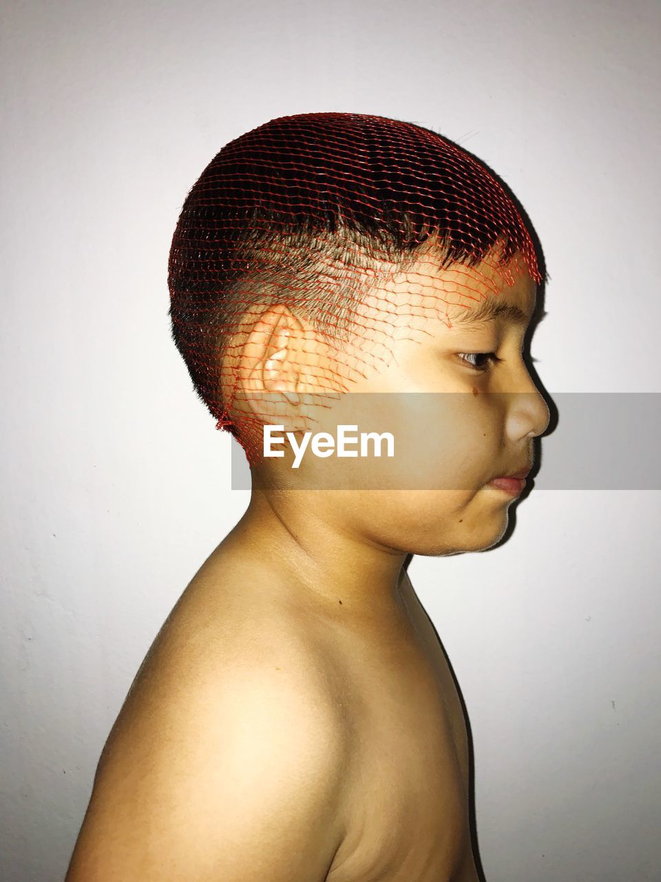Side view of shirtless boy wearing netting on head against white wall