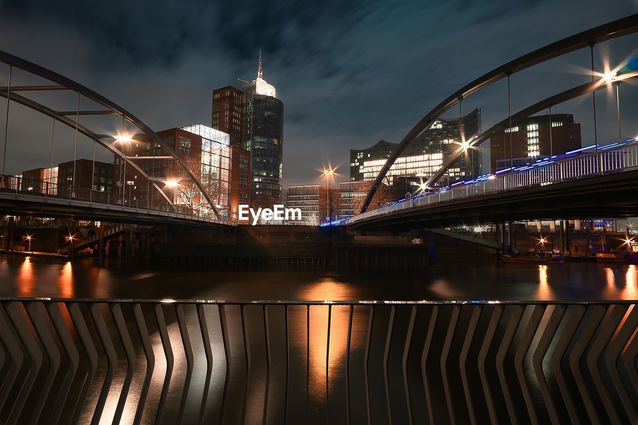 Illuminated bridge over river against buildings at night