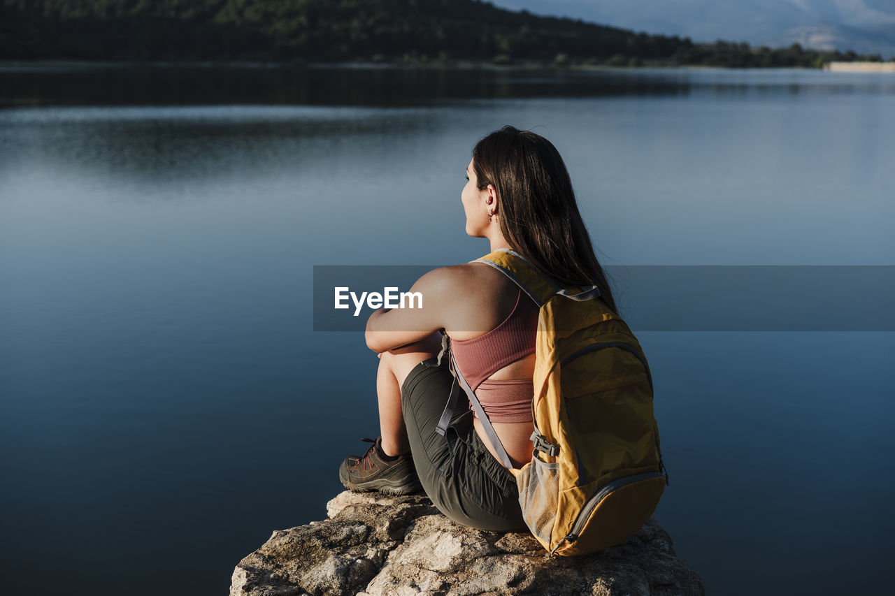 Young female backpacker with backpack sitting on rock by lake