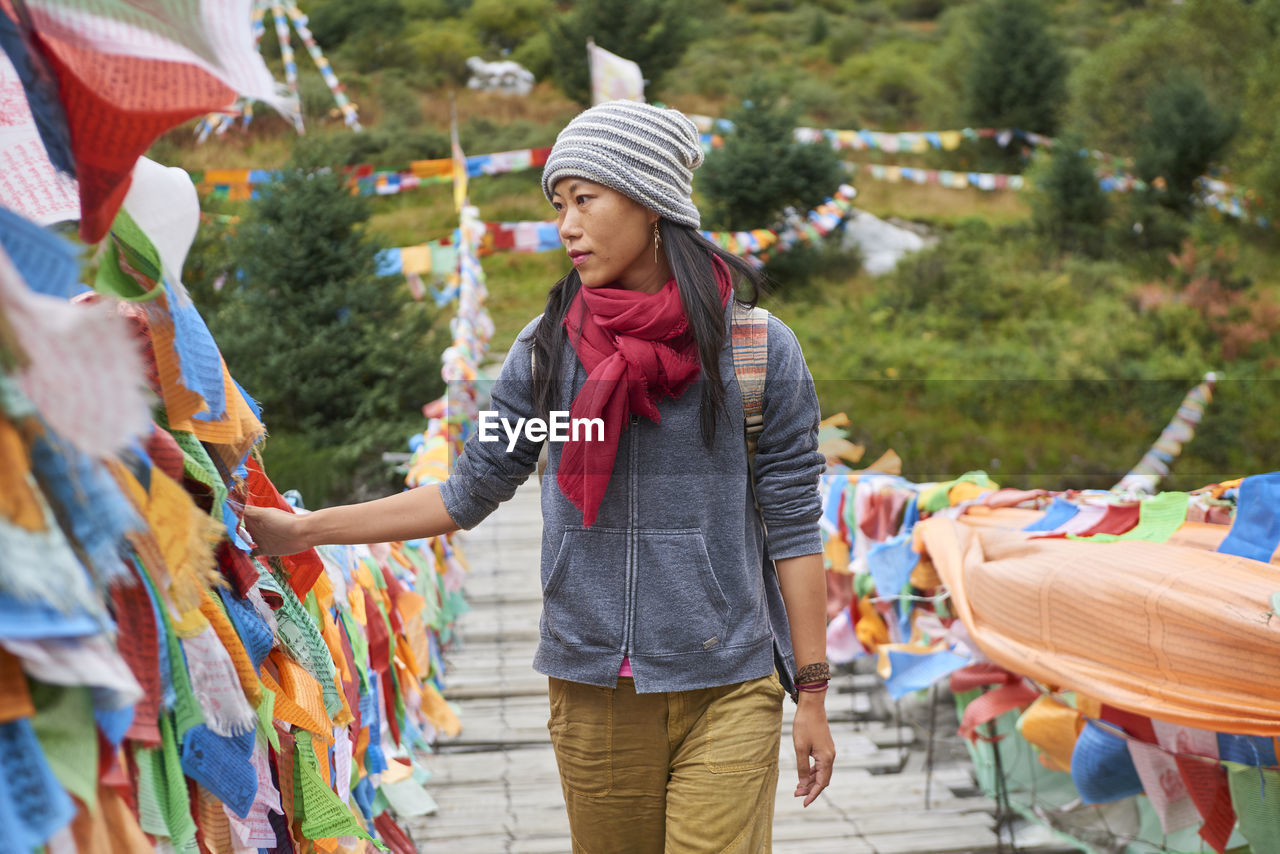 Young ethnic female tourist in warm clothing and hat looking away while crossing simple long narrow suspension wooden bridge decorated with bright multi colored pieces of fabric in daylight