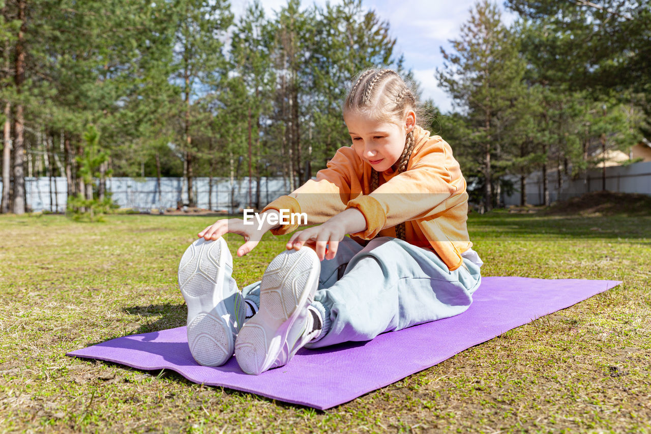 A teenage girl goes in for sports in nature, on a sunny summer day sits on a sports mat, stretches. 
