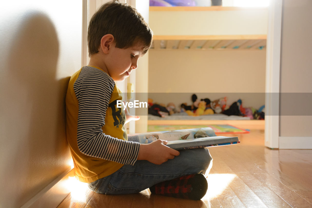 Side view of boy reading book while sitting on floor at home