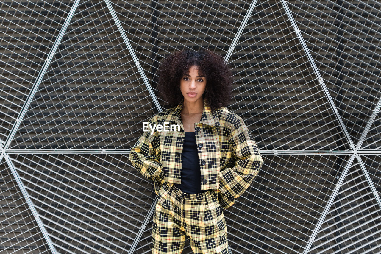 Young ethnic female in trendy checkered suit with hands behind back looking at camera in daytime