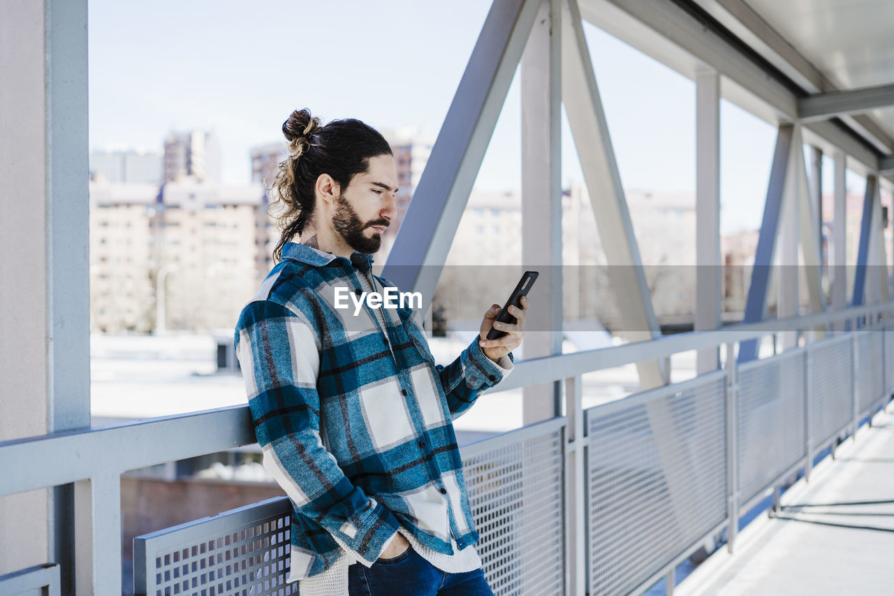 Young man using mobile phone while leaning on railing of bridge