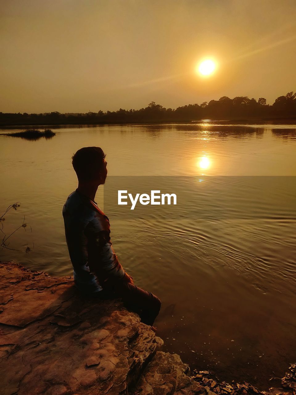 Side view of silhouette man sitting by lake against sky during sunset