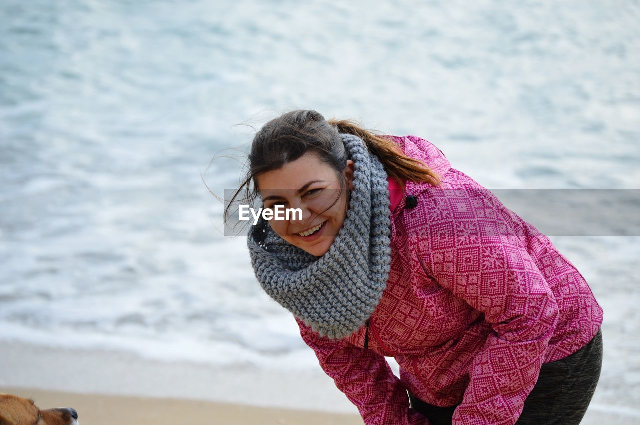 Portrait of smiling young woman wearing warm clothing while bending against sea at beach