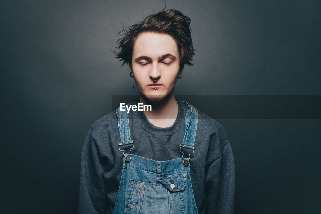 Young man with eyes closed wearing denim overalls over gray background