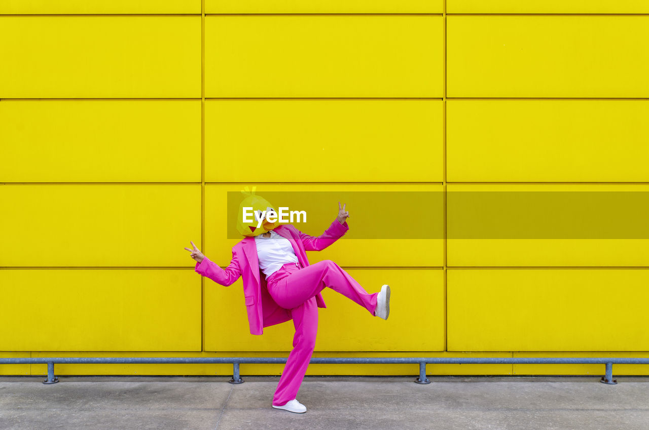 Woman wearing vibrant pink suit and bird mask making peace sign gestures while standing on one leg in front of yellow wall