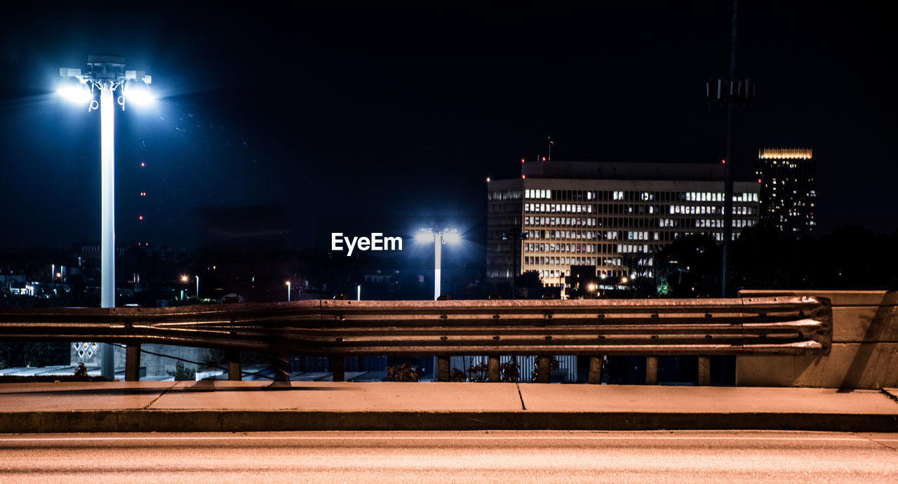 Illuminated street lights by bridge against buildings at night