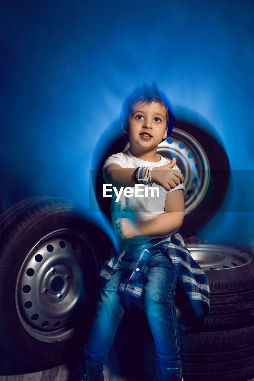 Boy in a white t shirt shirt and hat sits on a background of car wheels on a blue