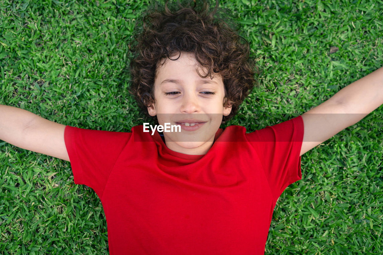 Cheerful boy with brown wavy hair, closed eyes and a red t-shirt lying on down on the grass