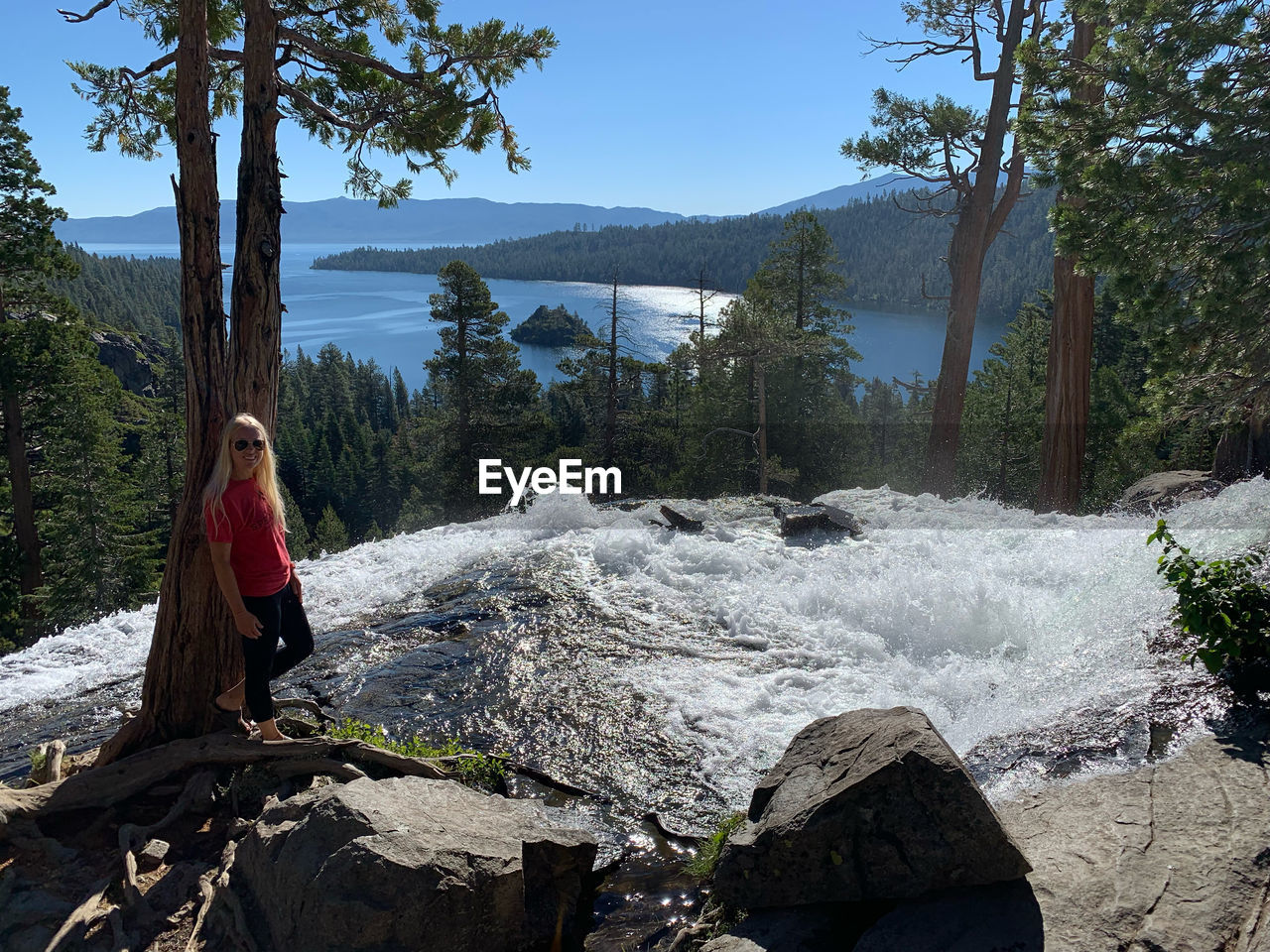 Front view of woman leaning against tree by lake and waterfall