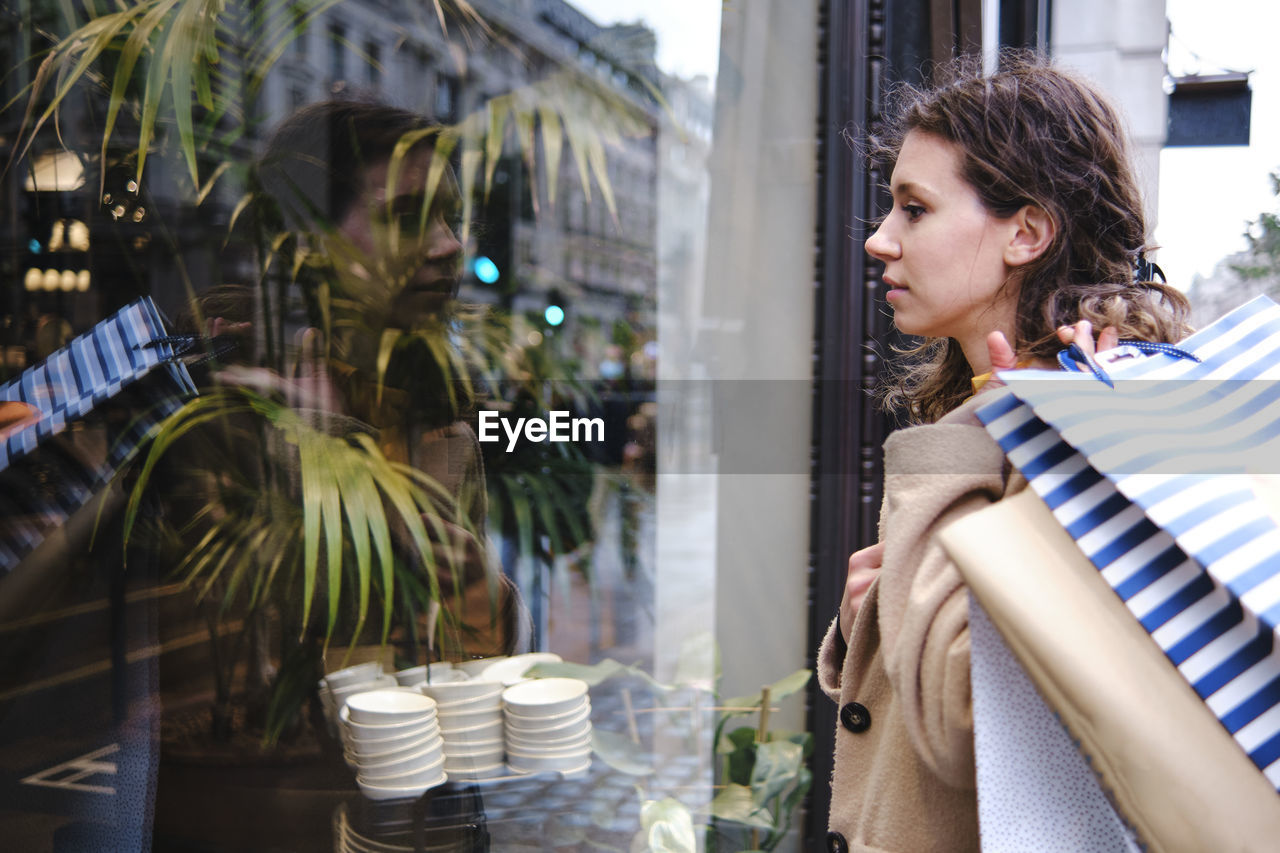 Woman looking through glass window of store while carrying shopping bags