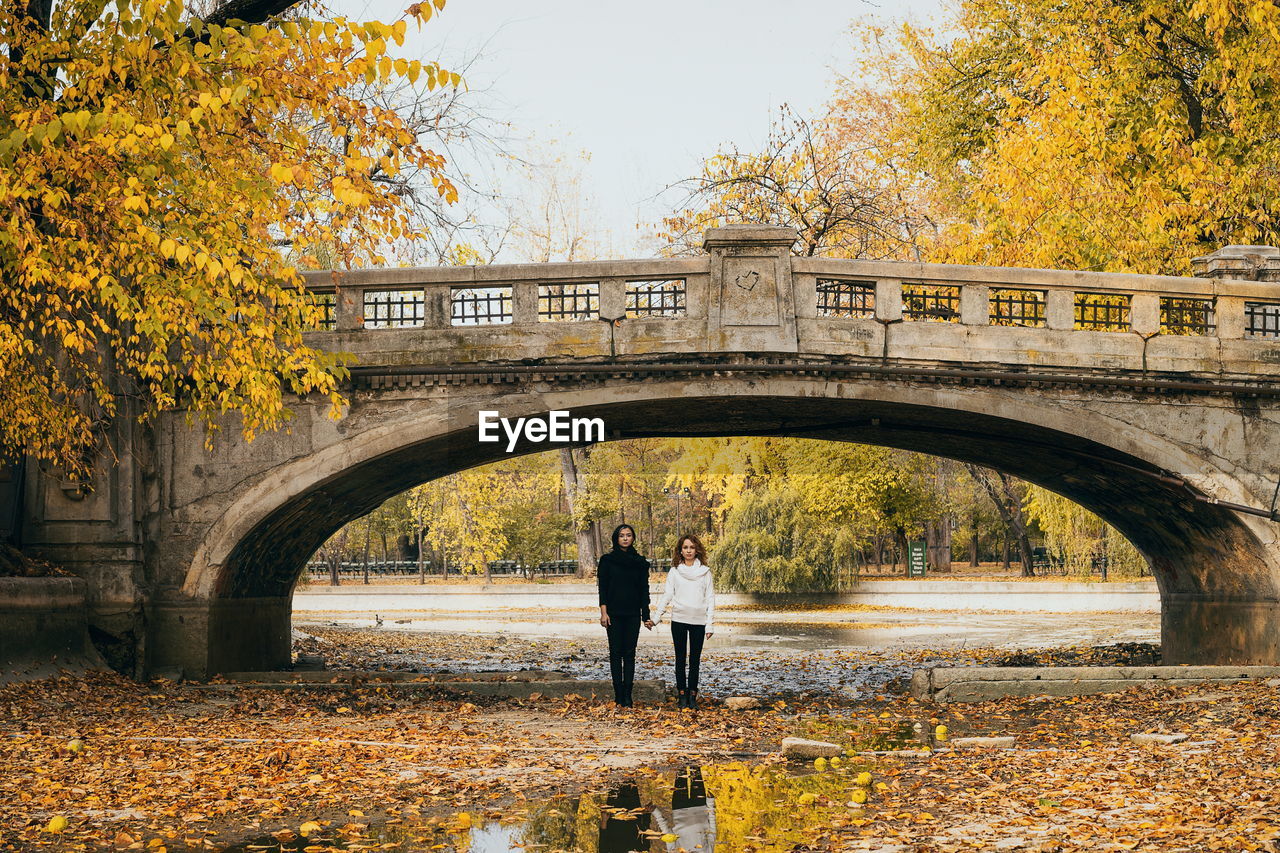 Lesbian couple holding hands while standing at arch bridge during autumn