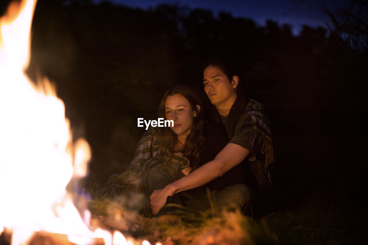 Couple wrapped in blanket sitting by campfire at night