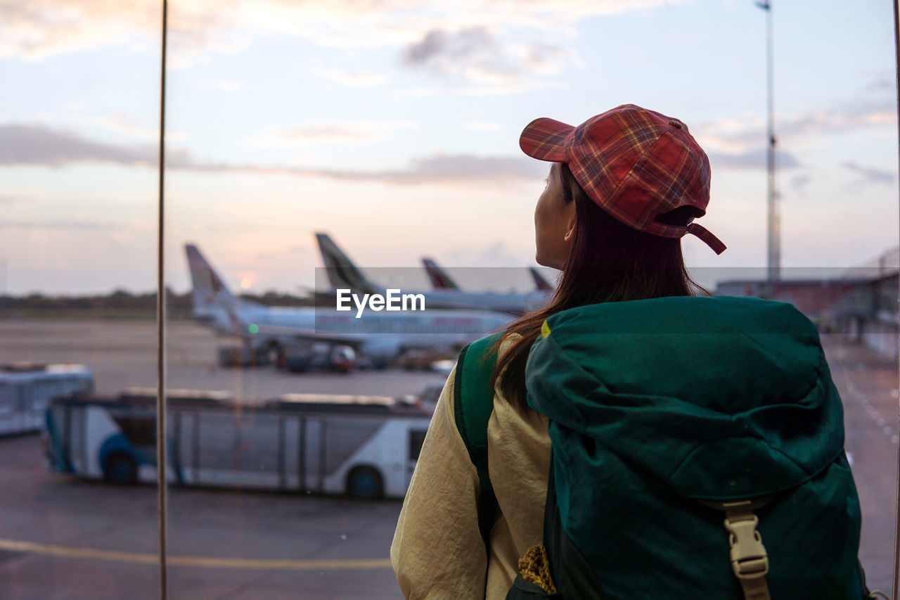 Back view of young asian female traveler with backpack standing near window in airport terminal and observing airplanes while waiting for flight in sri lanka