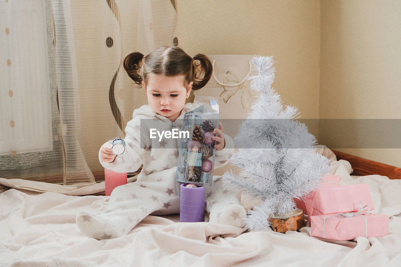 Full length of cute girl playing with christmas decoration while sitting on bed at home