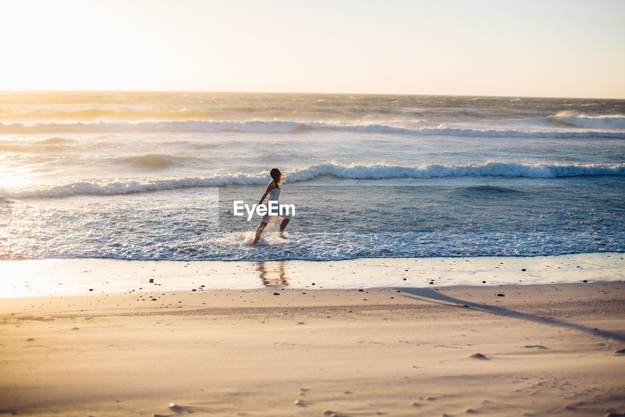 Young woman running at beach against clear sky during sunset